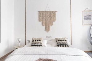 A modern bedroom with a bed, two patterned pillows, a macrame wall hanging, a small table with a vase, and a framed picture on the wall. - Banxia Inn (Hengdian Mingqing West Road) (Dongyang)