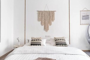 Un dormitorio moderno con una cama, dos almohadas estampadas, un colgador de macramé, una mesita con un florero y un cuadro en la pared.