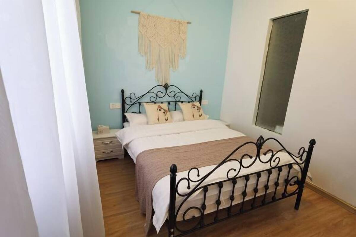 A bedroom with a bed, a nightstand, a wall-mounted macrame piece, and a window.