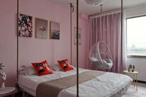 Un dormitorio con cama, dos almohadas, una silla colgante, una mesita pequeña y una ventana con cortinas.