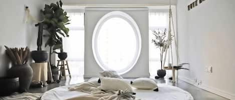 A minimalist room with a large round window, a bed with white linens, and a potted plant.