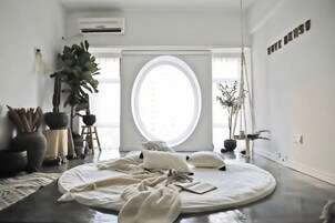 Una habitación minimalista con un ventanal redondo grande, una cama con ropa blanca y una planta en maceta.