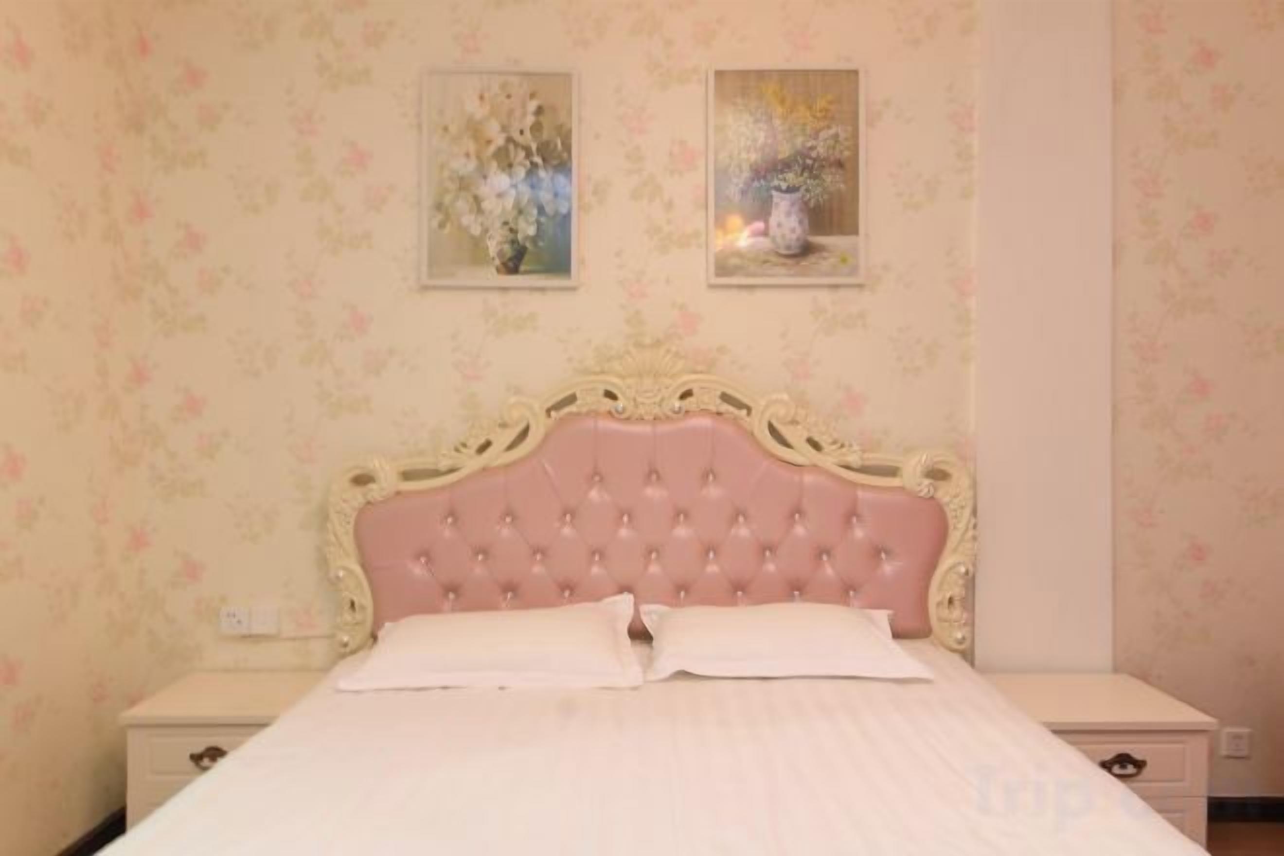 Un dormitorio con cabecera tapizada en rosa, colcha blanca y dos cuadros enmarcados con motivos florales en la pared.