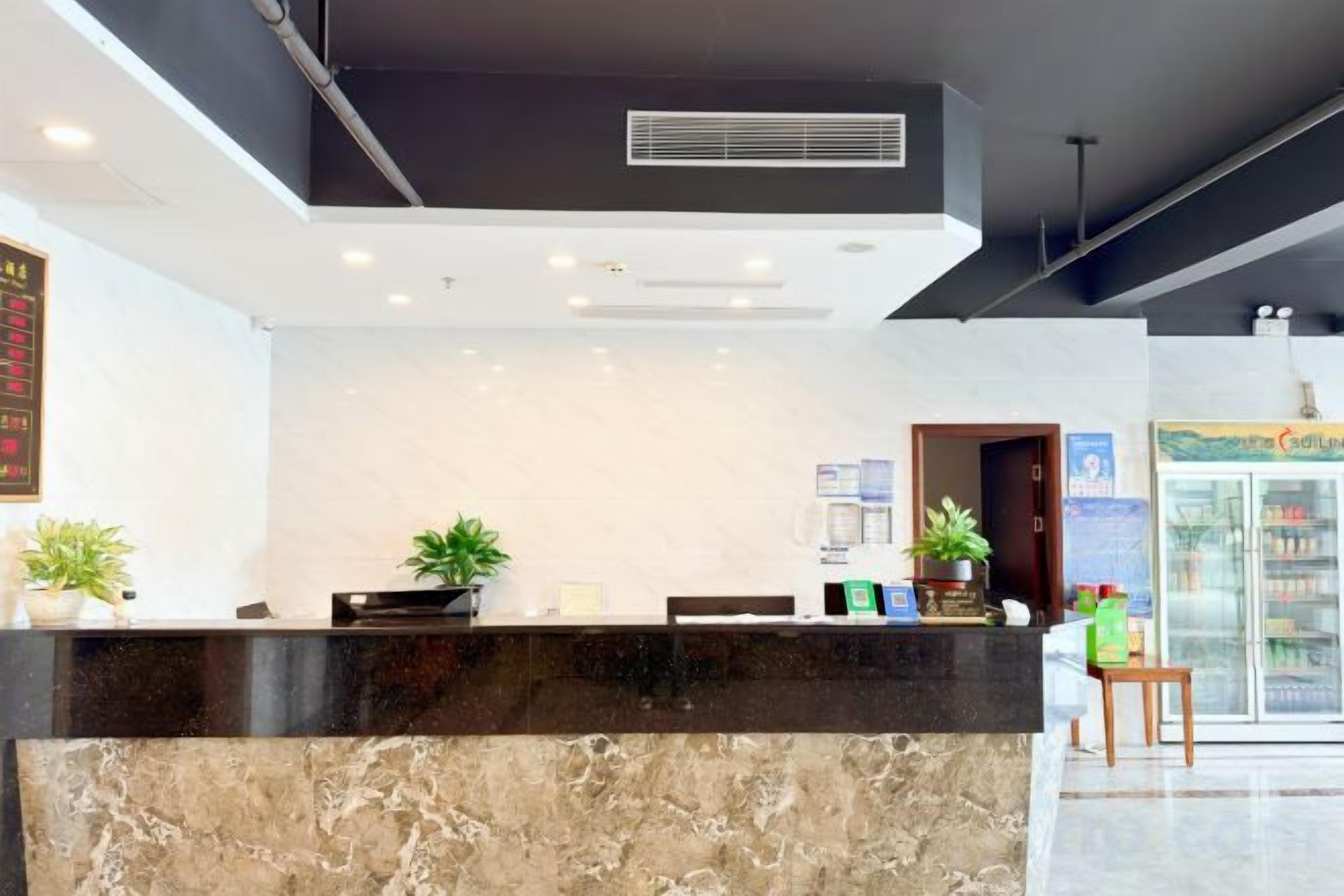 A hotel reception area with a marble counter, potted plants, and a refrigerator in the background.