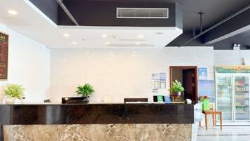 A hotel reception area with a marble counter, potted plants, and a refrigerator in the background.