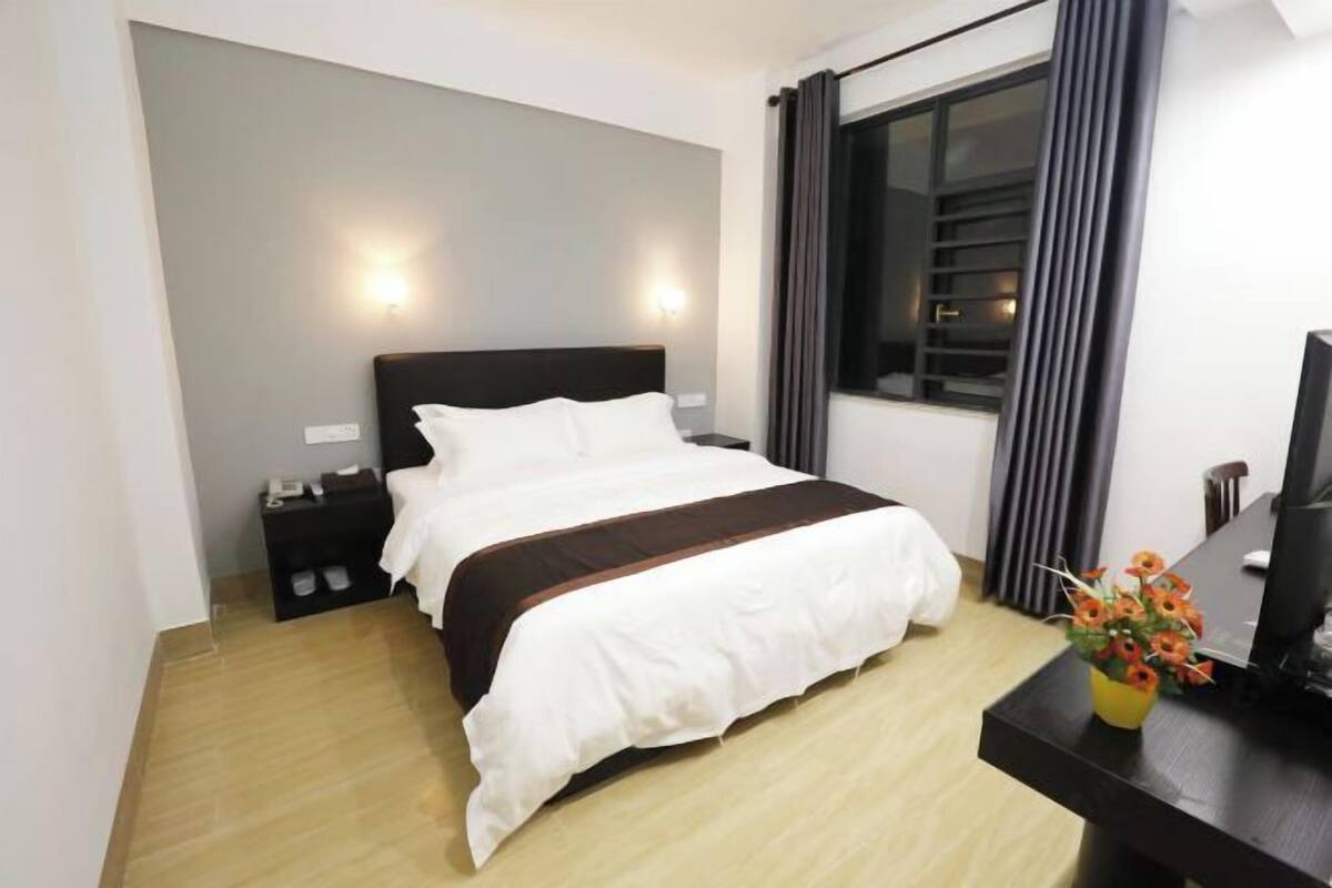 A hotel room with a bed, a desk with a computer, a nightstand, and a window with curtains.