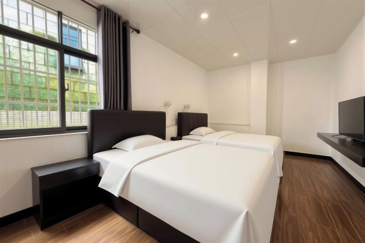 A hotel room with two beds, a TV, a window with curtains, and a bench.