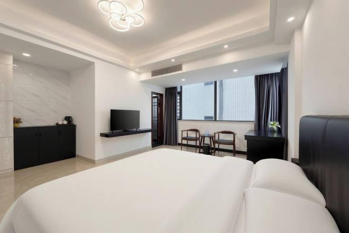 A modern hotel room with a large bed, a TV, a desk with a chair, and a window with curtains.