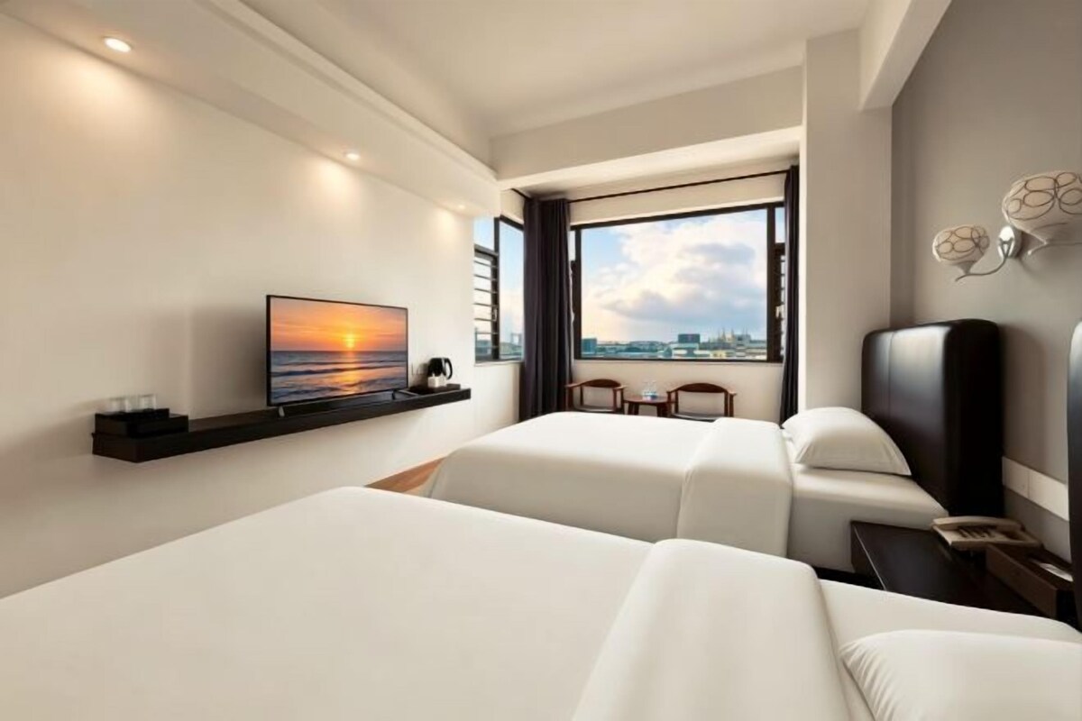 A hotel room with two beds, a TV, a window with a city view, and a small table.