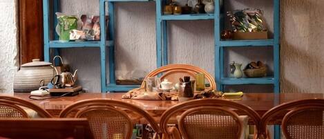A dining area with a wooden table and wicker chairs, a blue shelving unit with dishes, and a kitchen area with a kettle and pots.