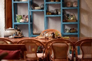 A dining area with a wooden table and wicker chairs, a blue shelving unit with dishes, and a kitchen area with a kettle and pots. - Huayutang Renwen Inn (Luoping)