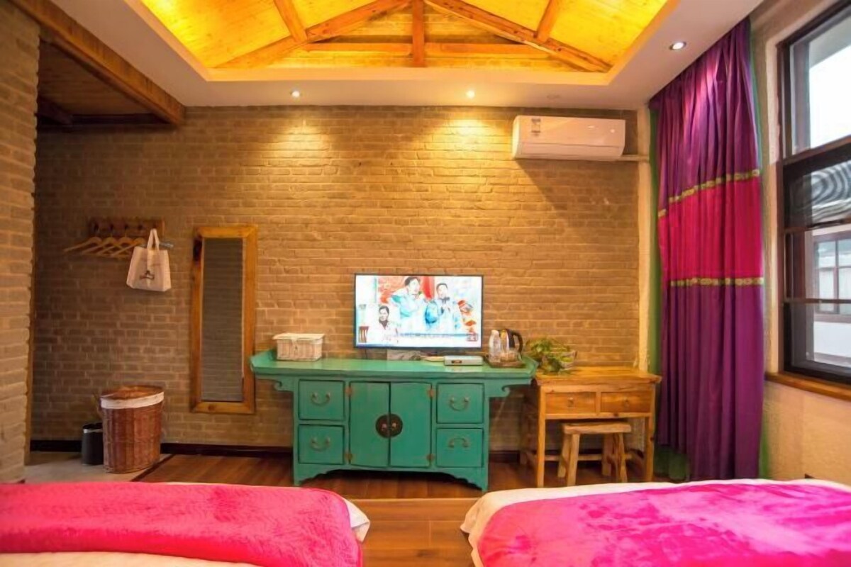 A room with a brick wall, a wooden ceiling, a television, a green dresser, a small table, and a window with purple curtains.