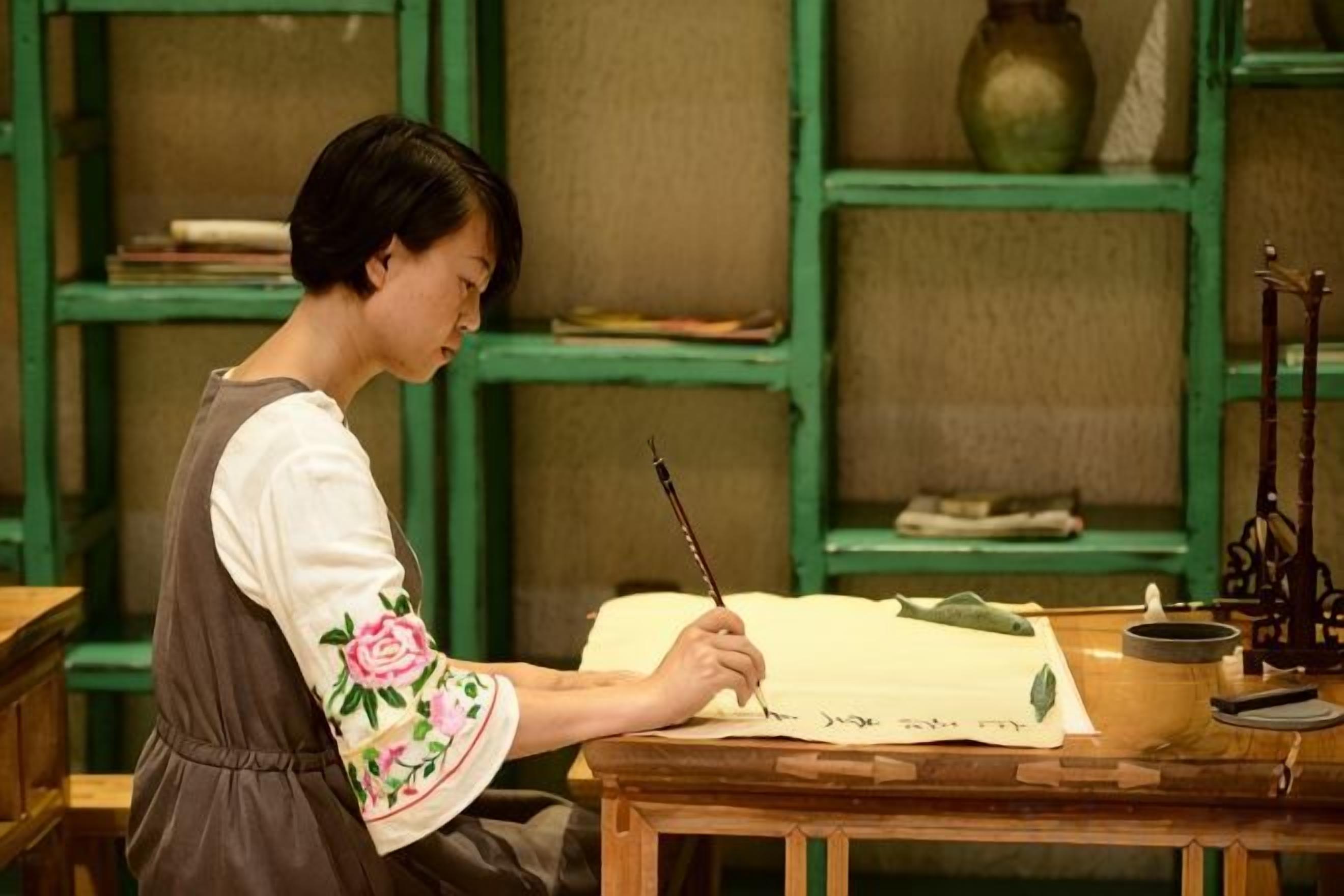 Una persona escribe sobre un papel grande en una mesa de madera, con una estantería verde al fondo.