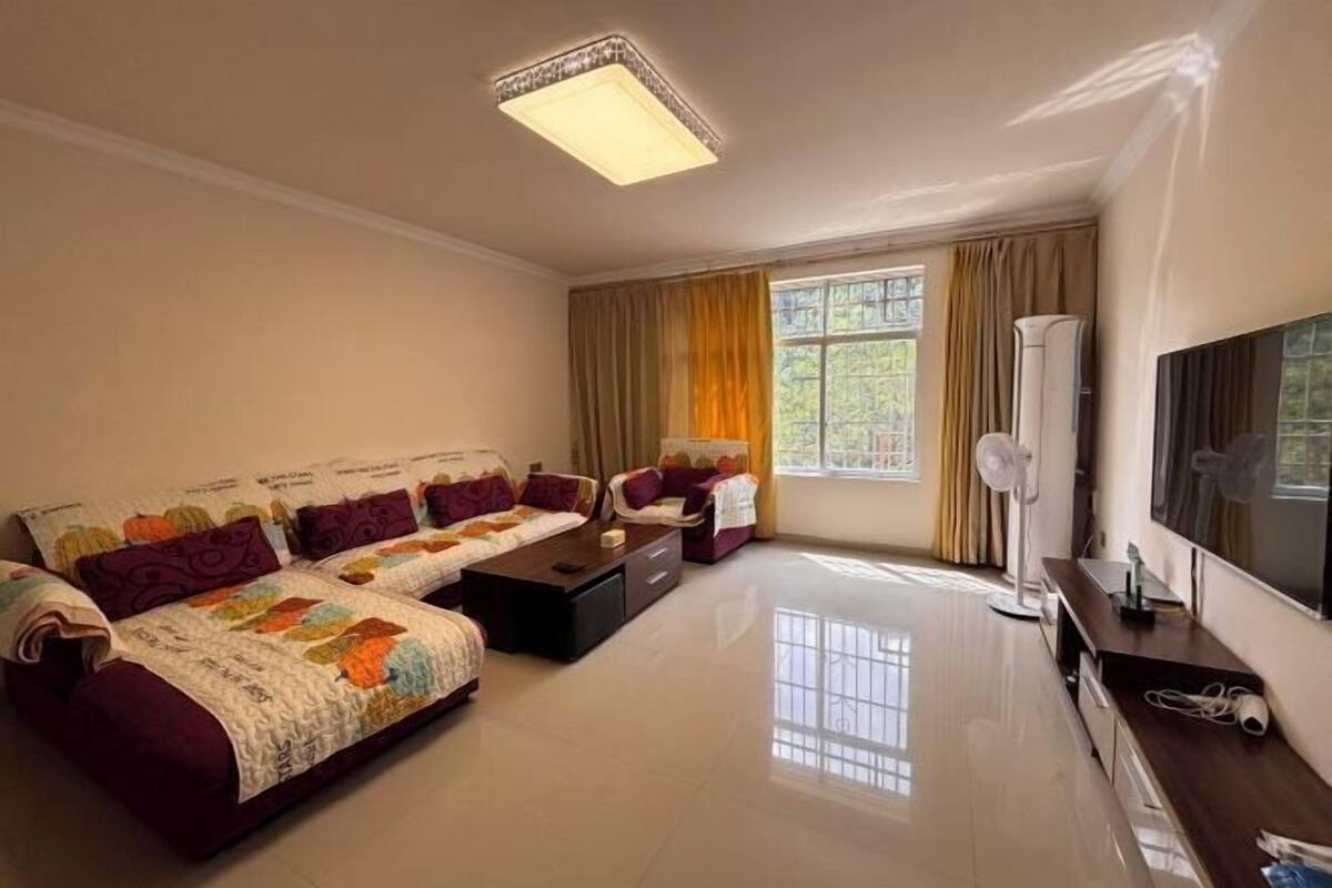 A spacious living room with a sofa set, a coffee table, a flat-screen TV, and a large window with curtains.