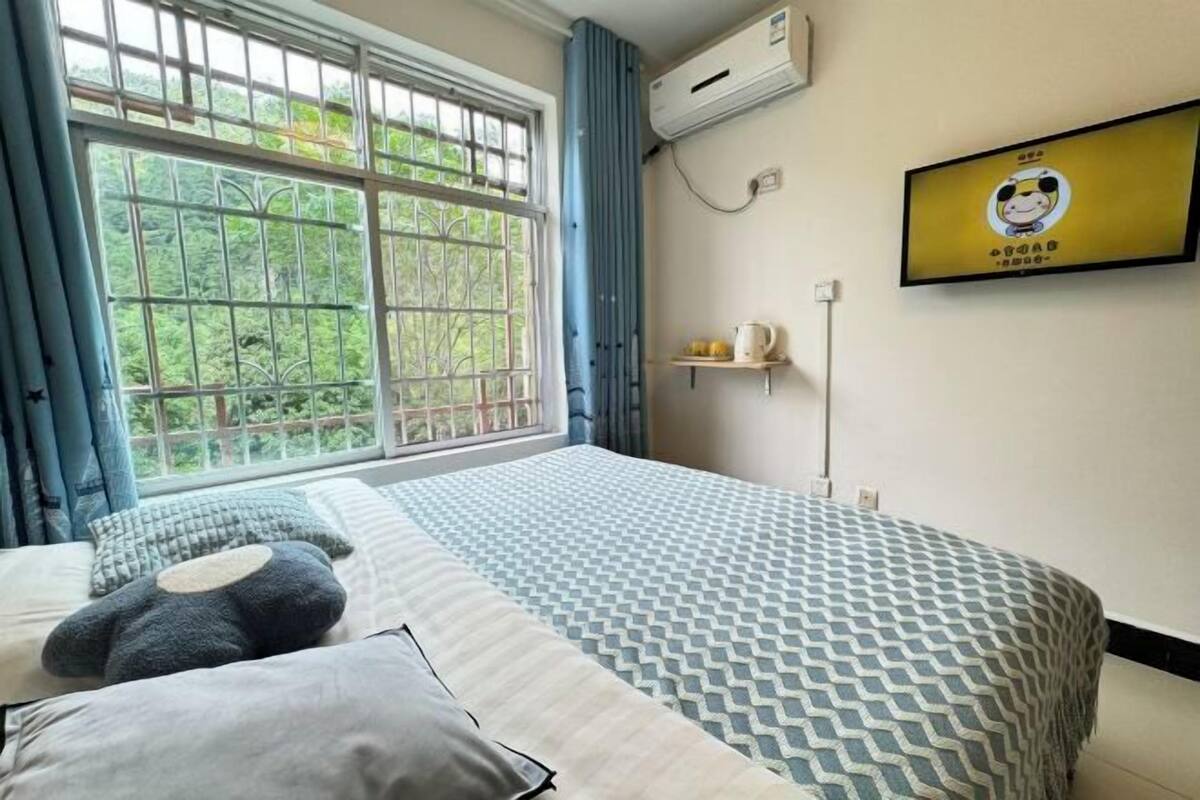 A bedroom with a large window, a bed with patterned bedding, a wall-mounted TV, and a shelf with a kettle.
