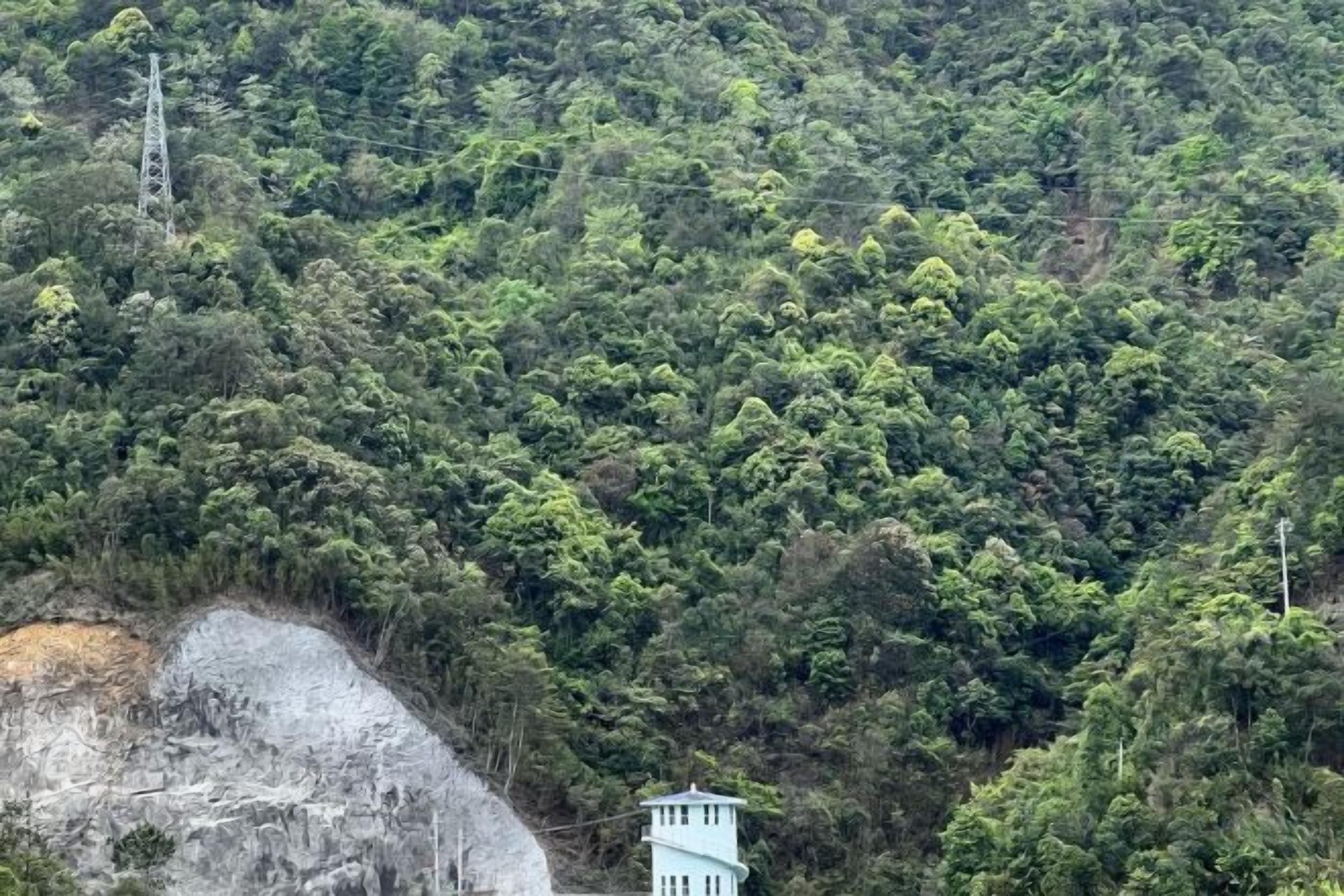 A building with a light blue exterior, surrounded by dense green forest and a rocky cliff.
