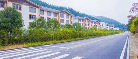 A road with a pedestrian crossing, buildings with multiple floors, and a hilly landscape in the background.