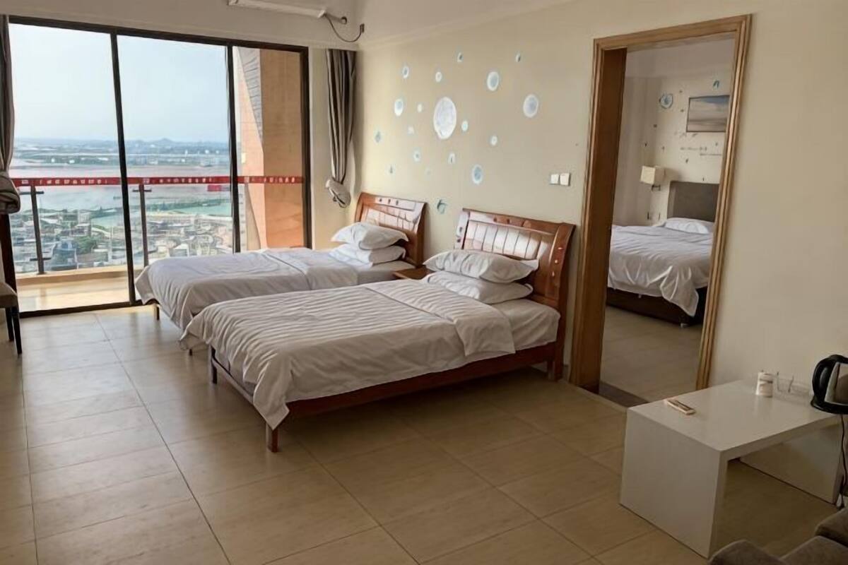 A hotel room with two beds, a desk, a large window with a city view, and a balcony.
