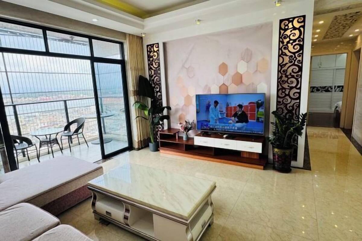 A modern living room with a large TV, a sofa, a coffee table, and a balcony with a view.
