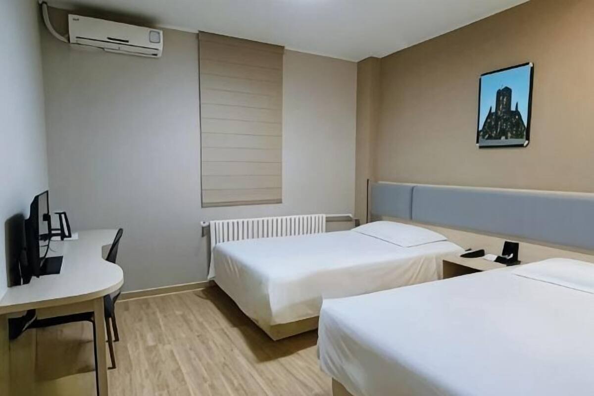 Habitación de hotel con dos camas individuales, un escritorio con silla, un radiador y un aire acondicionado instalado en la pared.