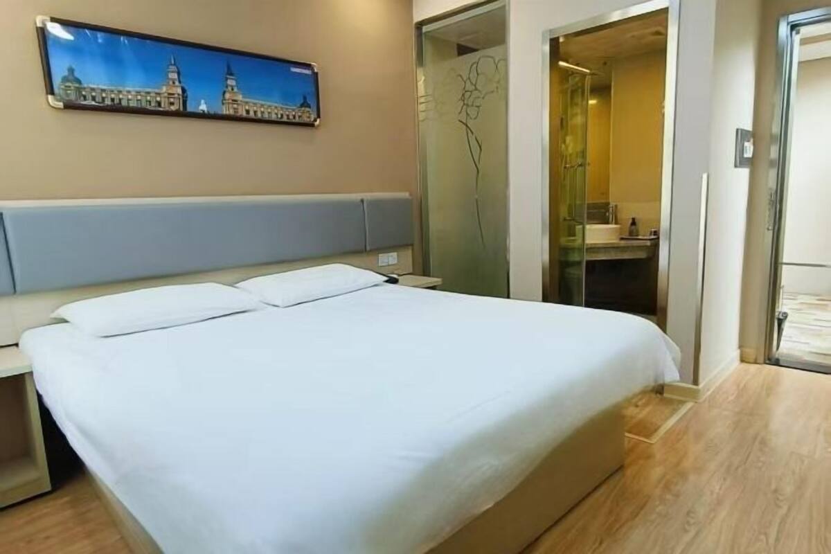 Una habitación de hotel con una cama grande, una mesita de noche, un televisor montado en la pared y una puerta de cristal que da a un baño.