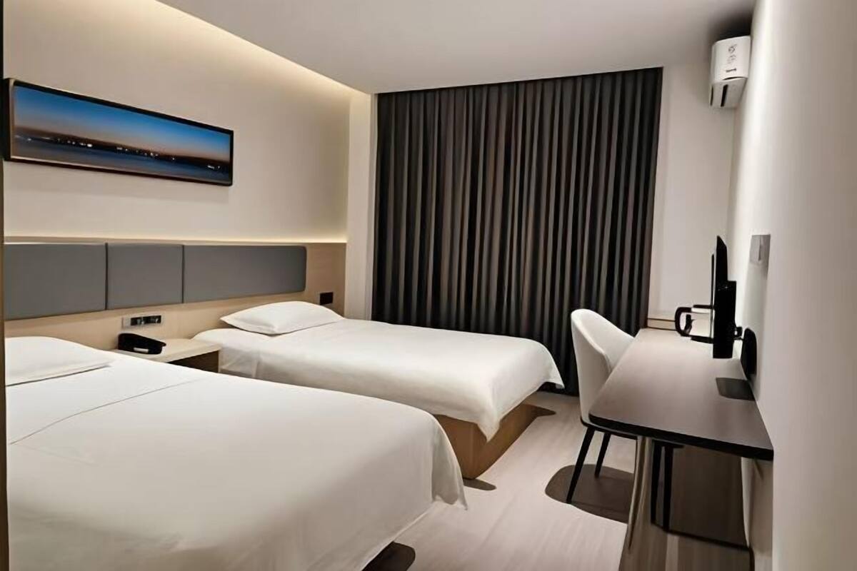 Habitación de hotel con dos camas, un escritorio, una silla, una cafetera y un ventanal grande con cortinas.
