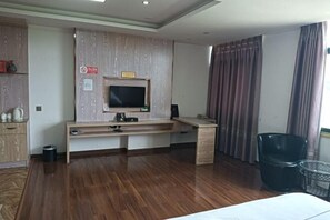 Habitación de hotel con piso de madera, un escritorio, un televisor fijado a un panel de madera, un cesto de basura y una ventana con cortinas.