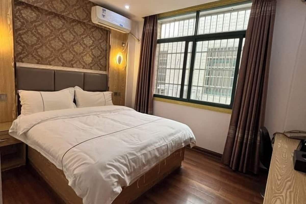 Une chambre avec un grand lit, une tête de lit en bois, un papier peint à motifs, une fenêtre avec des rideaux et un appareil de climatisation.