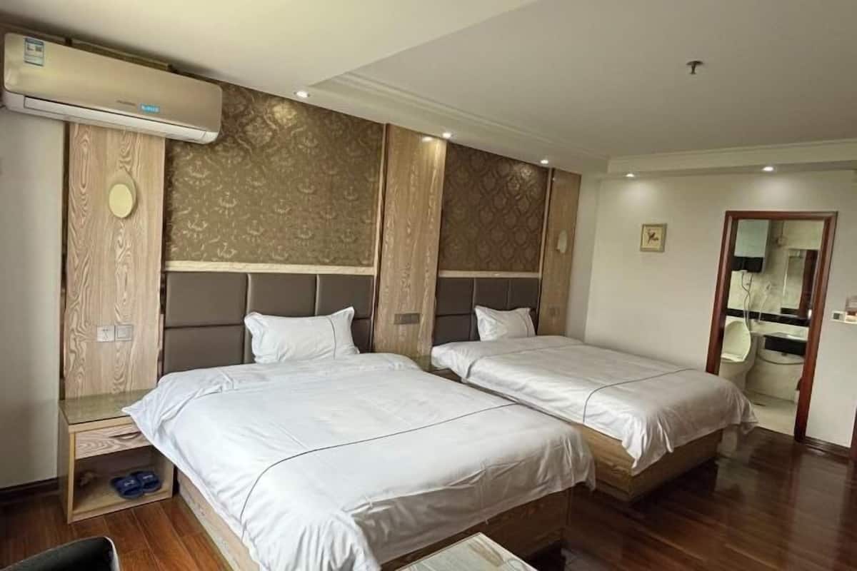 Une chambre d’hôtel avec deux lits, une armoire en bois, un appareil de climatisation et une salle de bain visible à travers une porte ouverte.