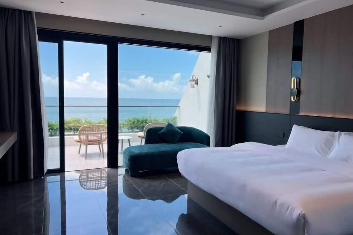 Habitación de hotel con cama, una silla, una mesita y vista al océano, además de un balcón.
