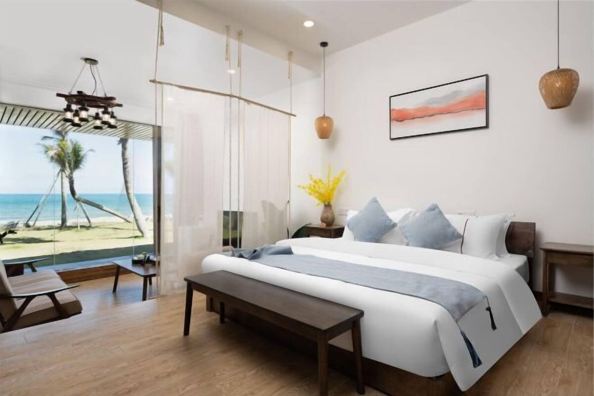 Un dormitorio moderno con una cama grande, un banco, una mesita de noche, una lámpara colgante y vista a la playa.