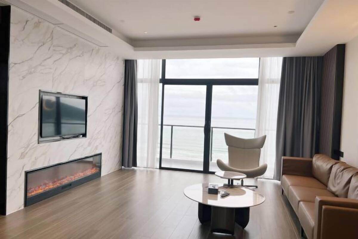 Una sala moderna con chimenea, televisor de pantalla plana, una mesa de centro redonda, un sofá de cuero y un ventanal con vistas al mar.