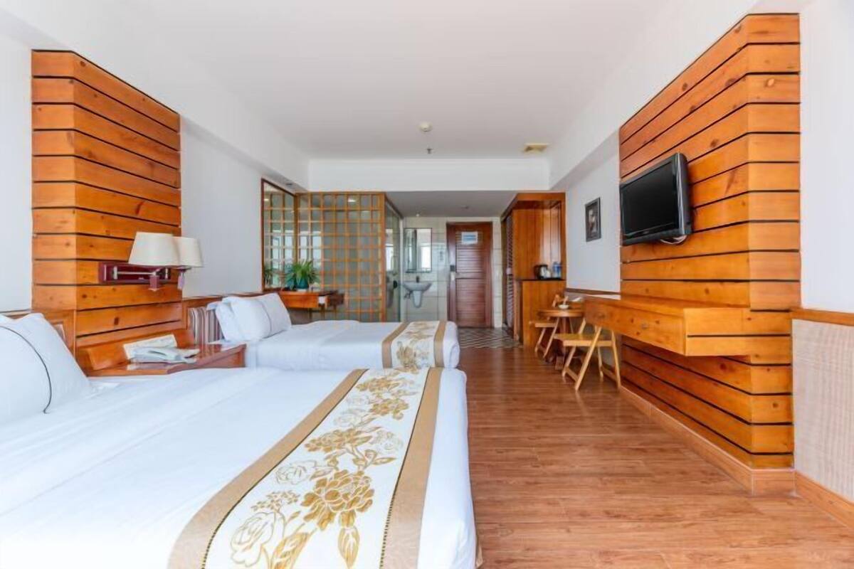 Habitación de hotel con dos camas, un televisor montado en la pared, un separador de madera y una zona de comedor.