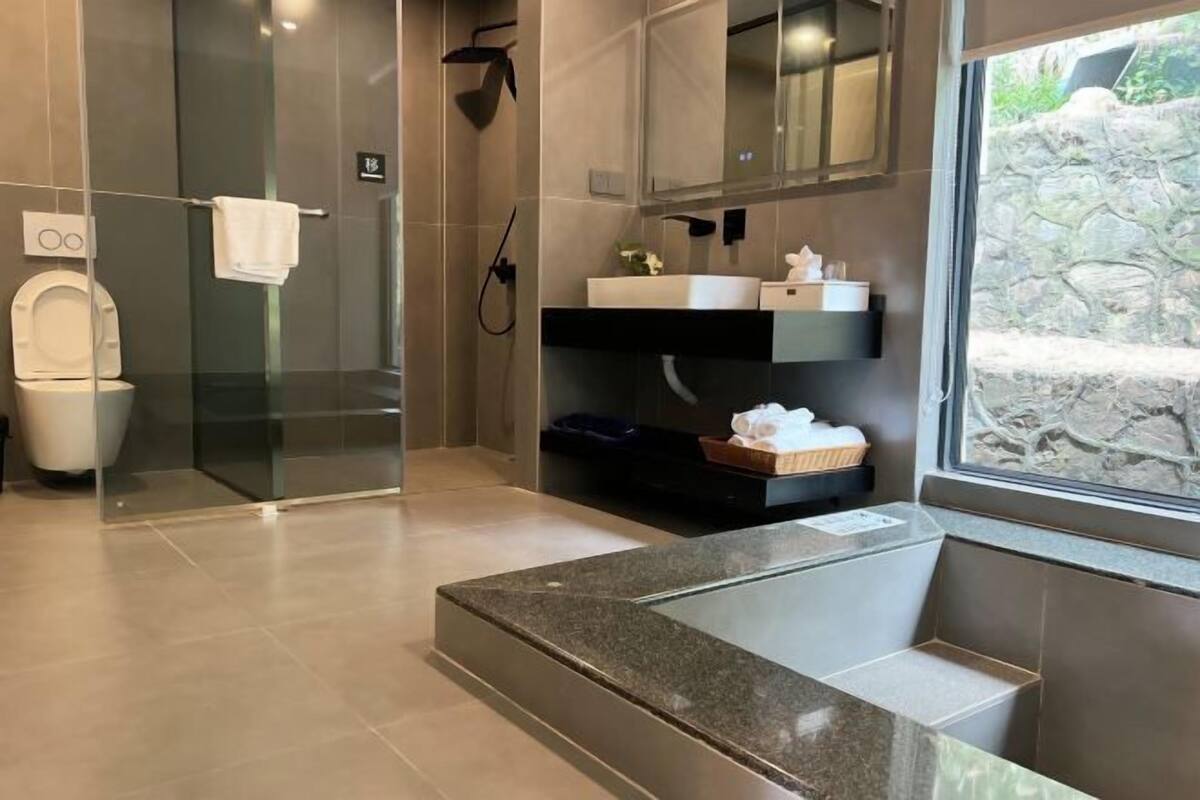 Un baño moderno con cabina de ducha de vidrio, lavamanos con encimera de mármol y pared de piedra visible a través de una ventana.