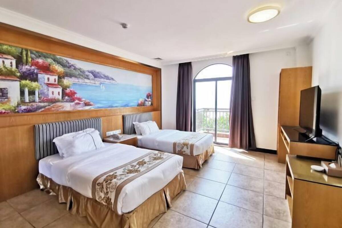 Habitación de hotel con dos camas, un cuadro de una escena costera, un televisor y un balcón con cortinas.
