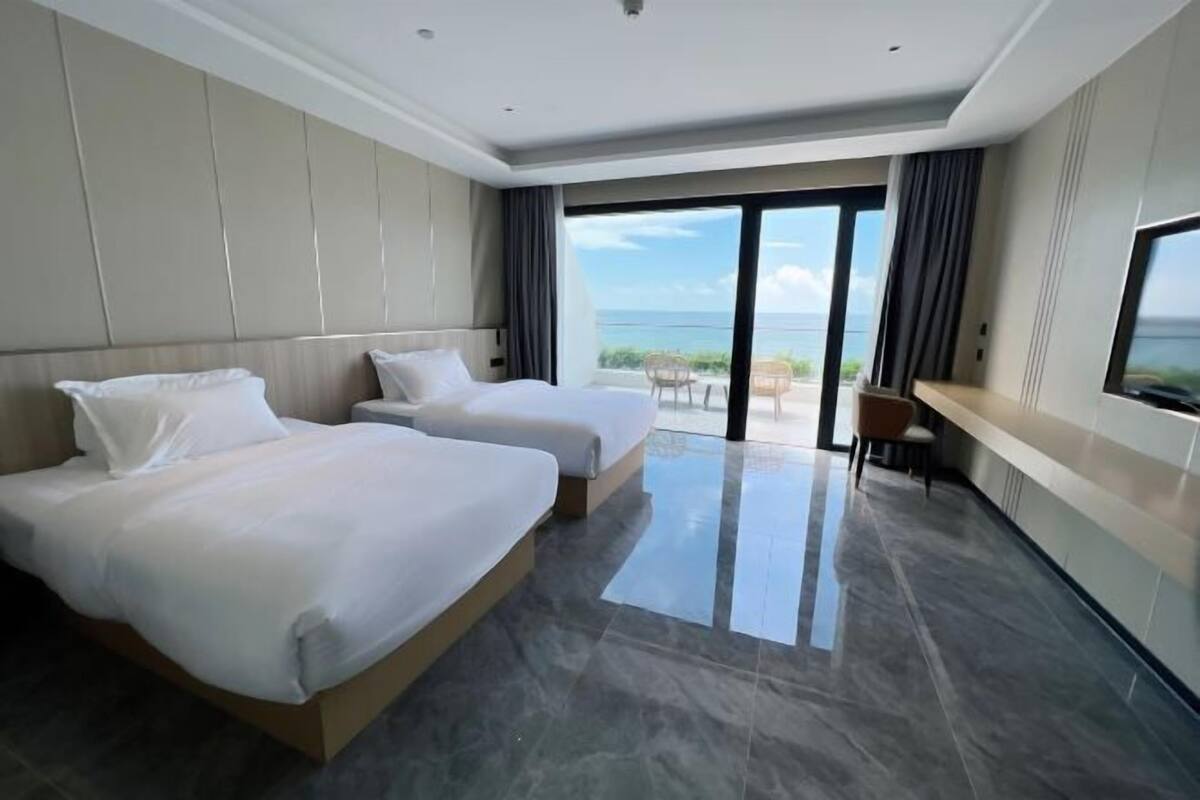 Una habitación de hotel moderna con dos camas, un ventanal con vistas al mar y un balcón con mobiliario exterior.