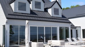 A modern house with a large glass sliding door, a covered patio with lounge chairs, and a view of the ocean.