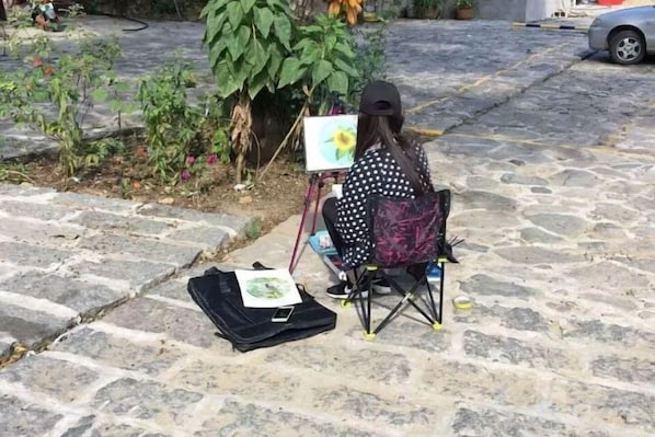 A person seated on a chair with a painting on a stand, a bag on the ground, and a plant nearby.