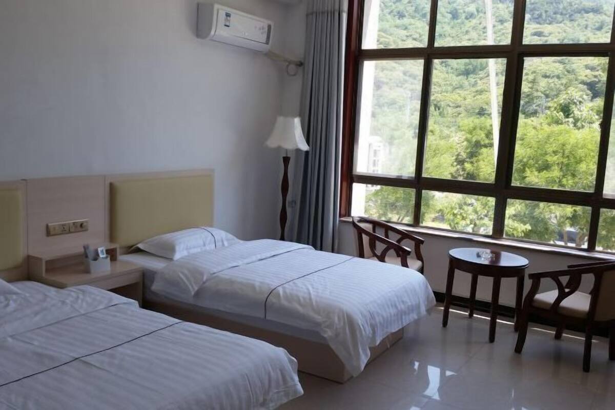 Habitación de hotel con dos camas, una mesita pequeña y vistas a la vegetación a través de amplios ventanales.