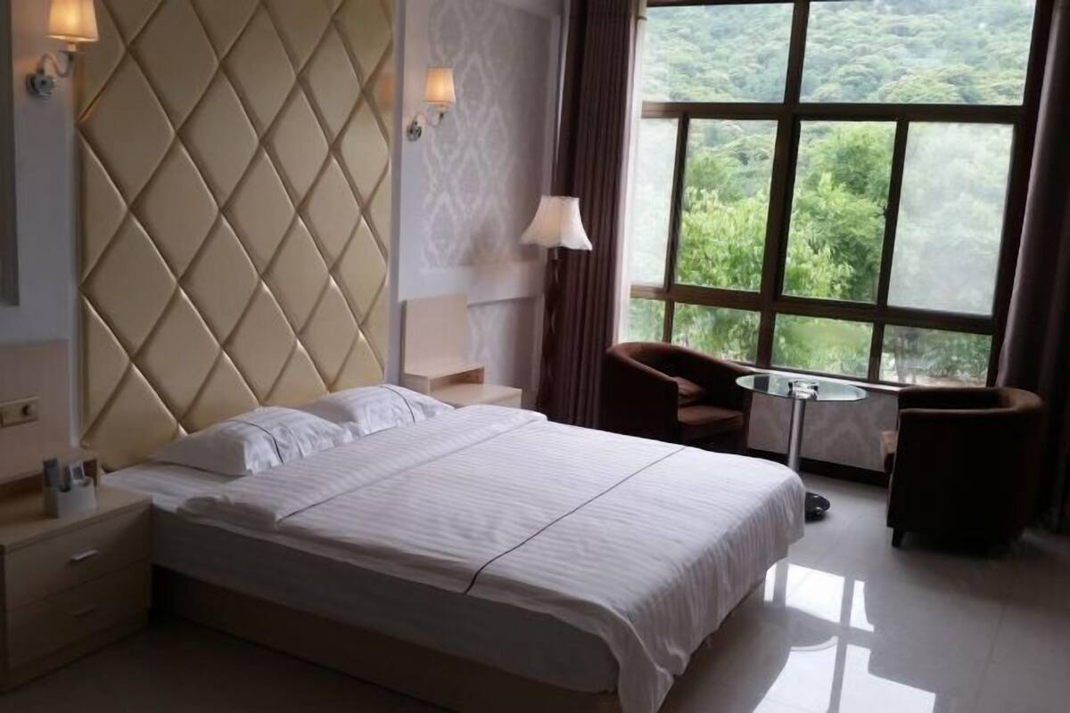 Habitación de hotel con una cama grande, una mesita de noche, una silla, una mesa y una ventana con vistas a zonas verdes.