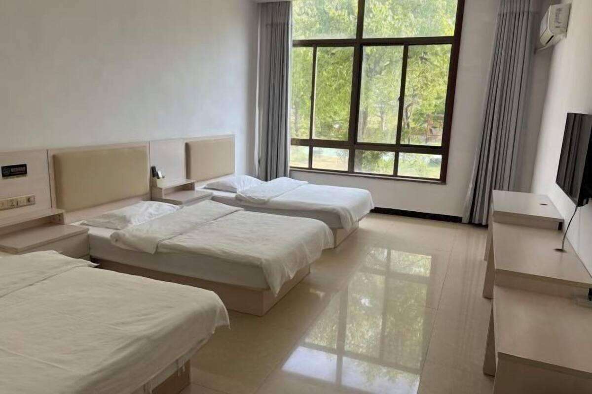 Una habitación de hotel con tres camas, un escritorio, un televisor y un ventanal grande con cortinas.