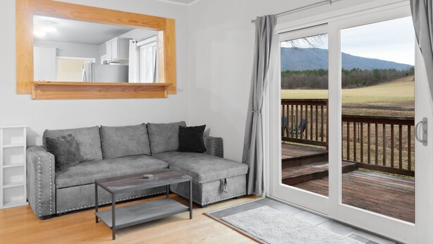 Ein modernes Wohnzimmer mit einer grauen Eckcouch, einem Couchtisch und einem großen Spiegel. Eine Glastür öffnet sich zu einer Terrasse mit Blick auf eine Wiese und Berge.