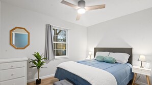 Ein Schlafzimmer mit Bett, Nachttischen, Deckenventilator, Fenster, Spiegel, Kommode und einer Topfpflanze.