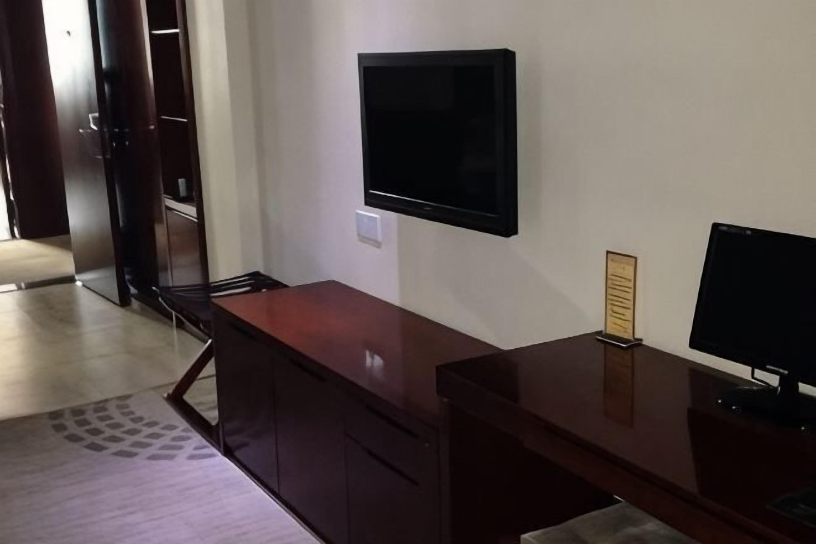 Una habitación con un escritorio de madera, un televisor de pantalla plana en la pared, un armario pequeño y una puerta que da a otra habitación.