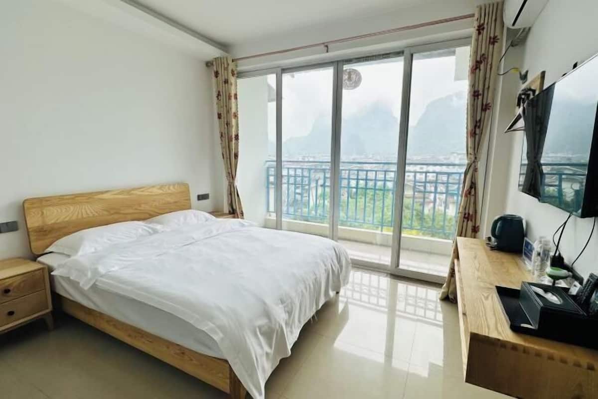 A bedroom with a wooden bed, a desk with a computer, and a large window with a view of a balcony and mountains.