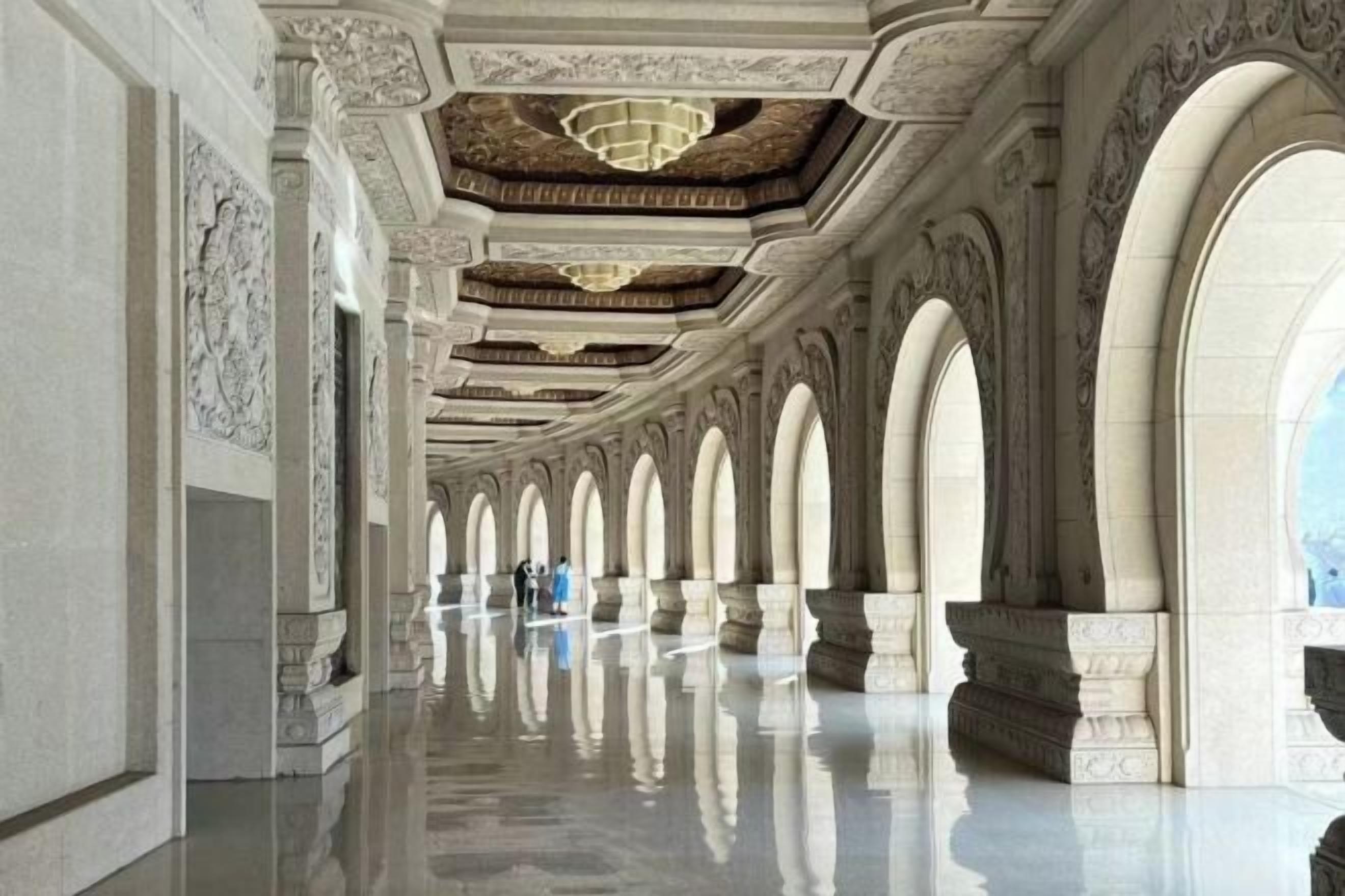 A corridor with arches and a reflective floor, leading to a large open space.