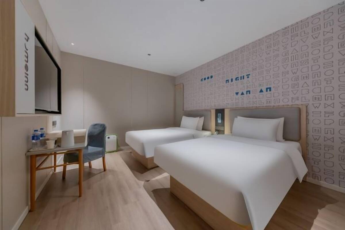 A hotel room with two beds, a desk, and a chair. There is a wall-mounted TV and a patterned wall.