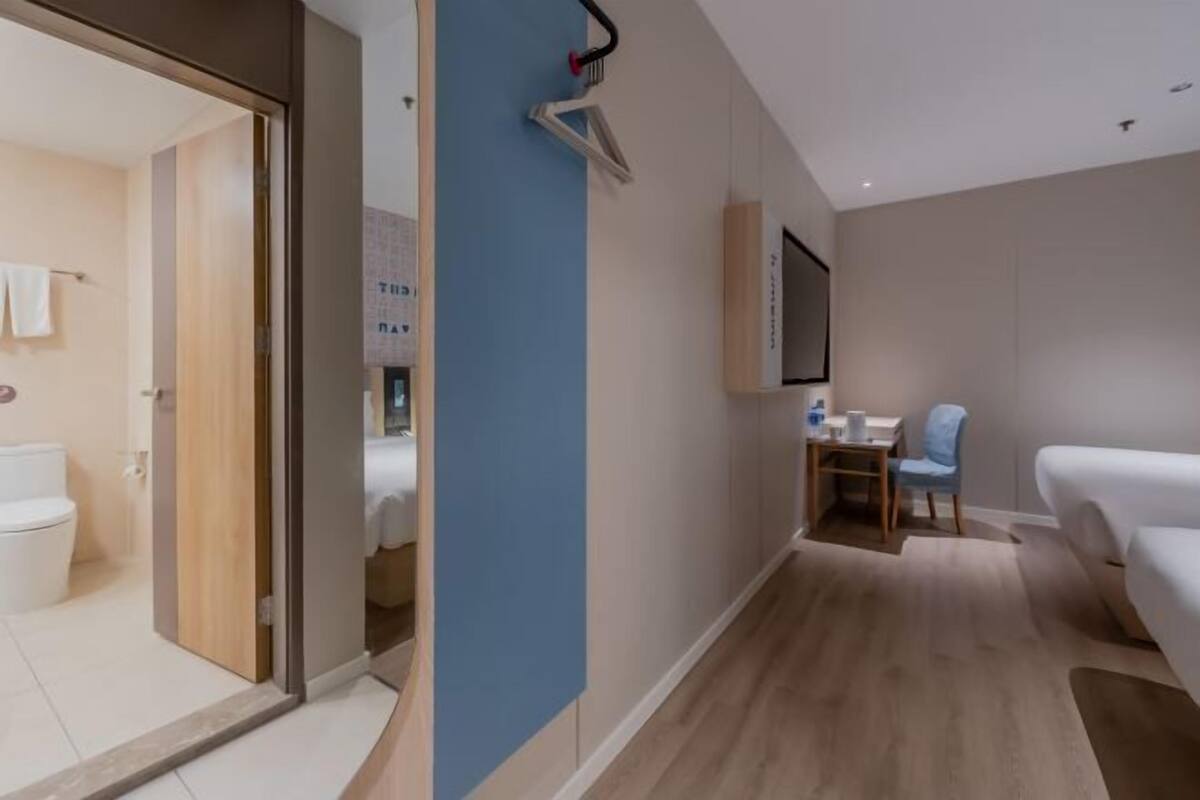 A modern hotel room with a wooden floor, a bed, a desk with a chair, a TV mounted on the wall, and a bathroom visible through an open door.