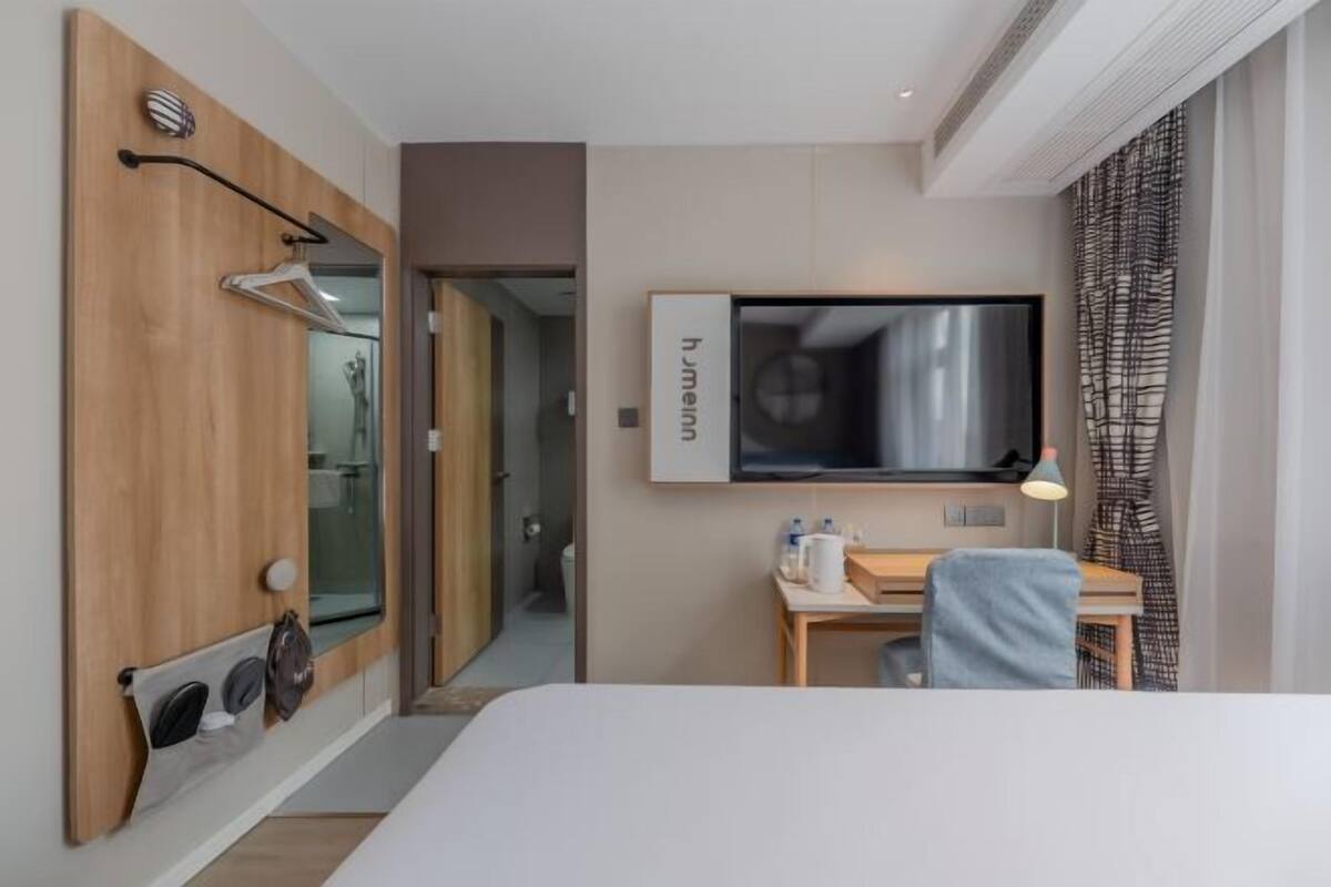 A hotel room with a bed, a desk with a chair, a television mounted on the wall, and a bathroom visible through an open door.