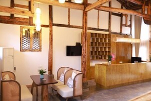 Uma área de recepção com balcão de madeira, sala de estar com poltronas estofadas e uma televisão na parede.